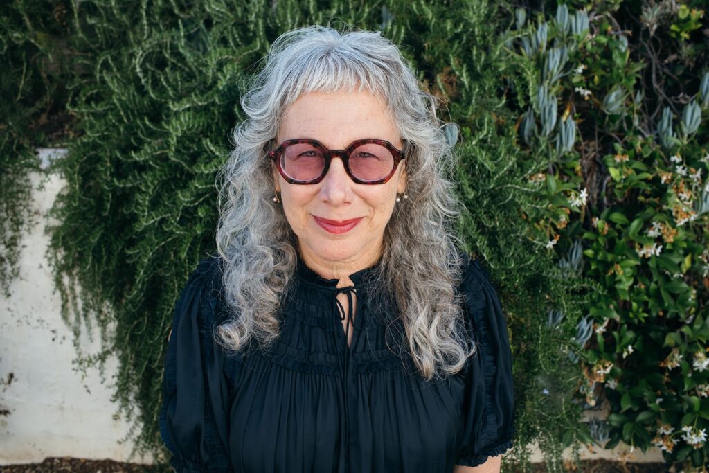 Portrait of designer Louise Sandhaus, smiling and looking at the camera against a backdrop of greenery.