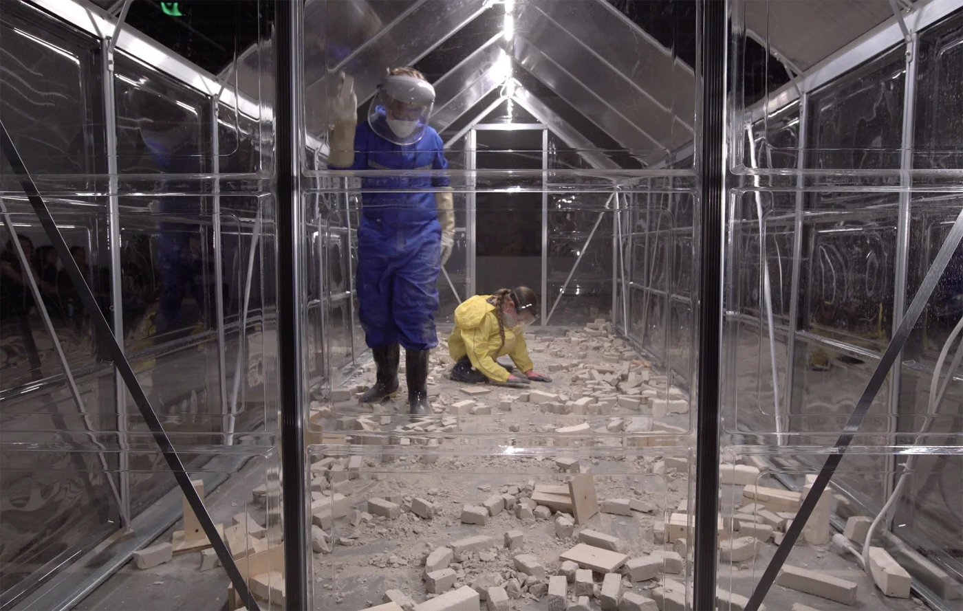 Art installation with two people wearing hazard suits in a transparent structure with debris scattered on the floow.