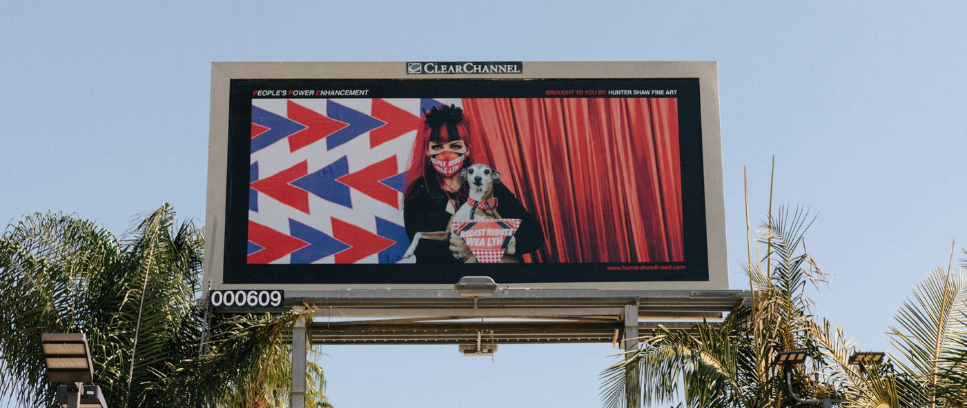 Billboard against a blue LA sky featuring a woman wearing a surgical mask with the words “redistribute wealth.” She holds a small dog and appears against a red, white, and blue backdrop.