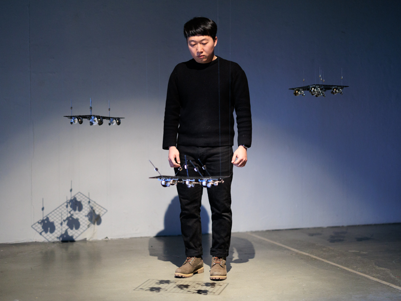 A person standing among three flat objects suspended from the ceiling in a dramatically lit gallery. 