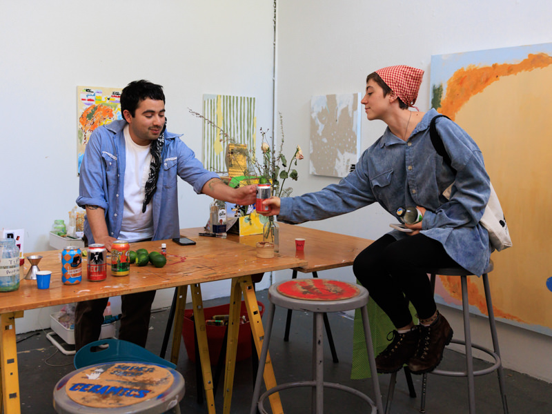 Two people at a makeshift bar in a small art studio passing a beverage