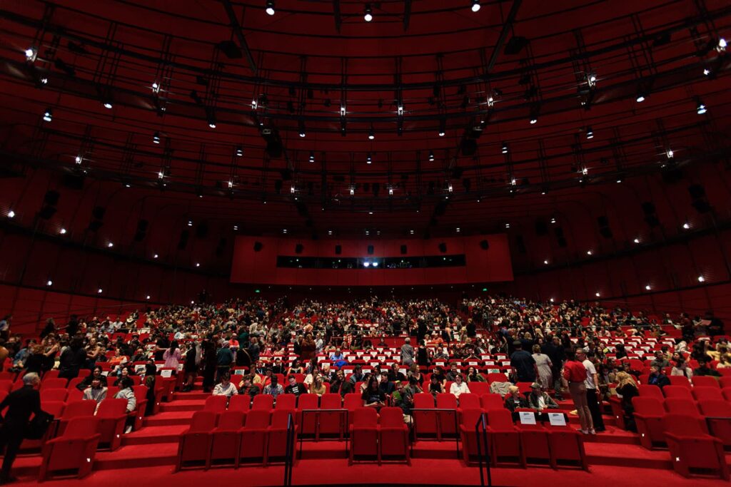 An audience seated in a glamorous theater with red carpets and velvet seats 