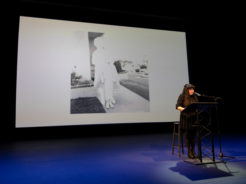 A person stands at a podium on a darkened stage with a black-and-white image on a screen behind her.