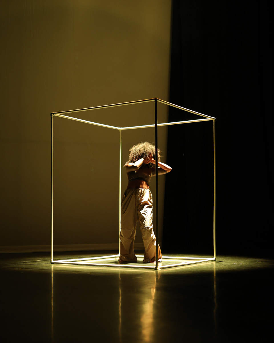 A dancer in loose cargo pants and curly hair stands in a single spotlight, inside a gold cube frame