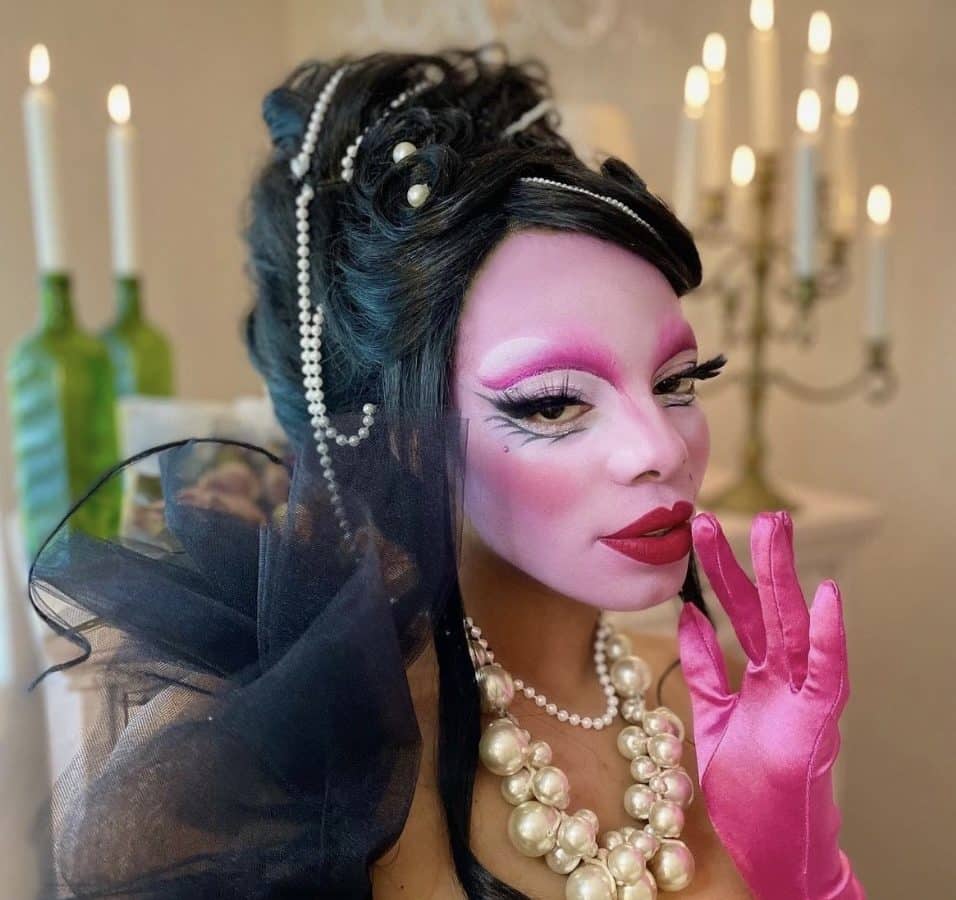 Person in elaborate pink makeup looks at the camera, raising a pink gloved finger to her mouth. 