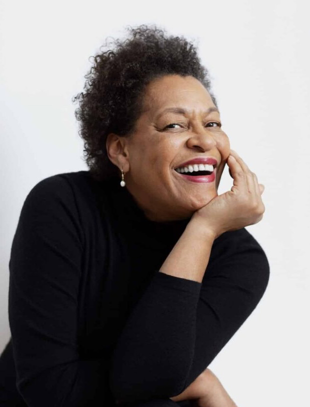 Photographic portrait of artist Carrie Mae Weems, laughing and dressed in black against a white background