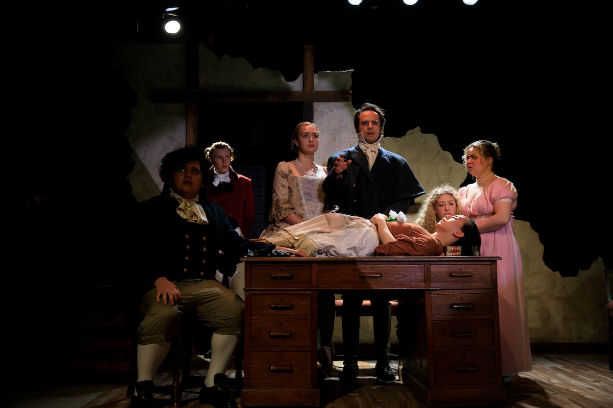 Actors in period costumes on a stage gather around a woman laid out on a desk.