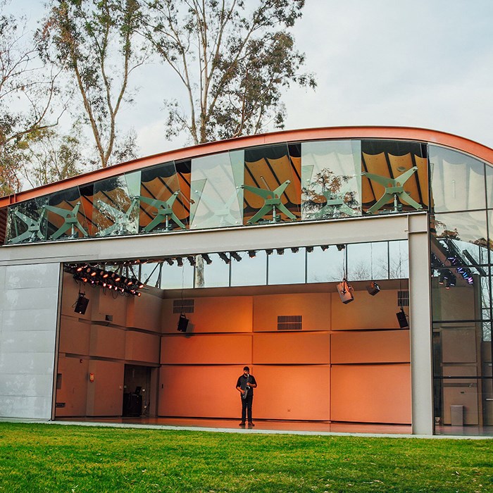 A lone performer plays a saxophone, standing in the middle of an empty stage in a futuristic bandshell with a lawn in front and trees and sky in the background.