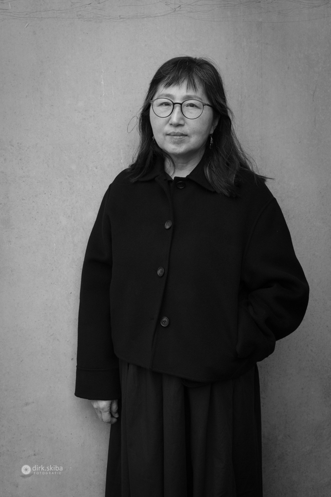 Black-and-white portrait of a woman in a dark coat with one hand in her pocket, standing against a plain background.