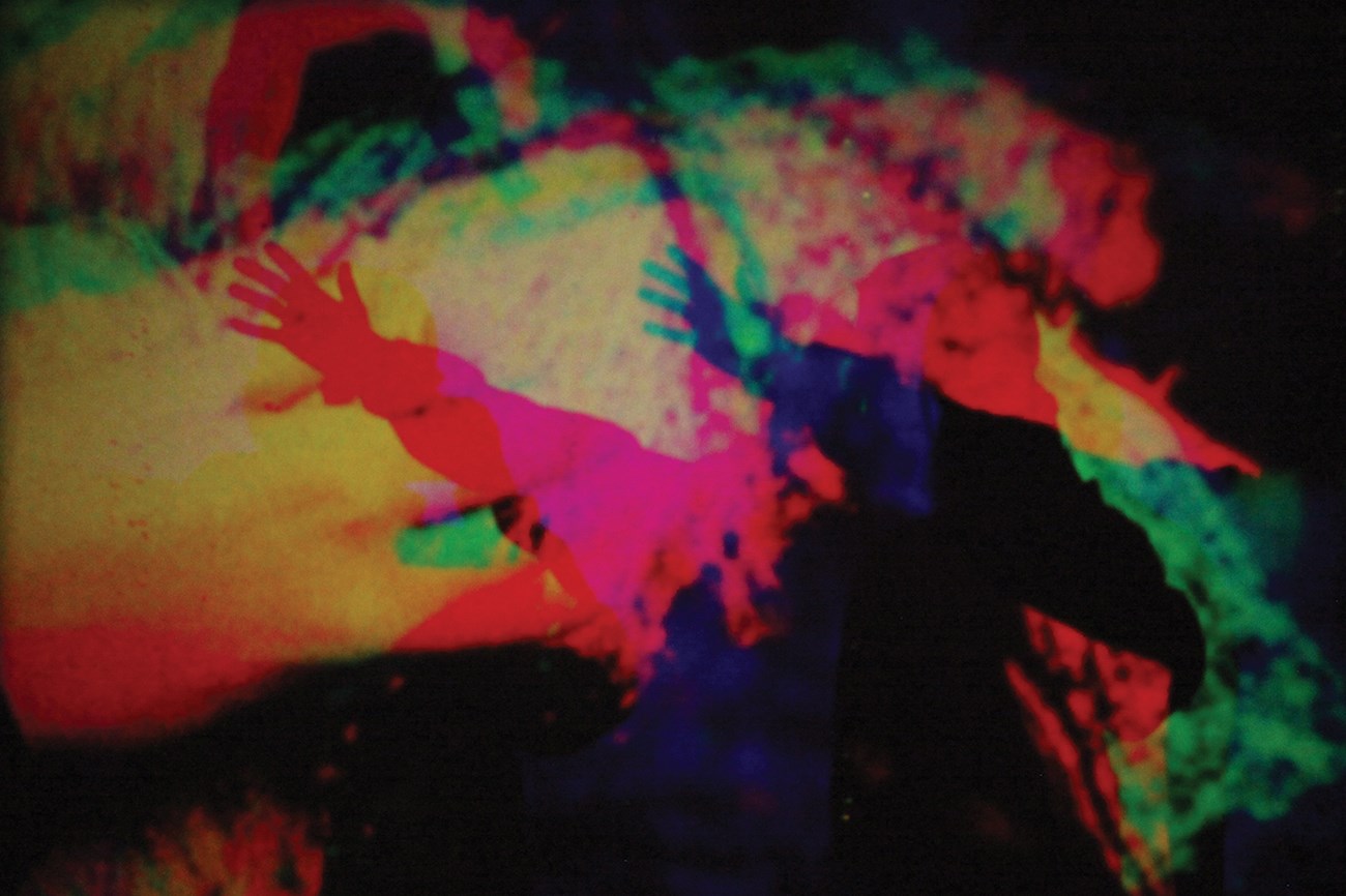 Brightly colored abstract image from an art film installation with silhouette of a figure with an outstretched hand. 