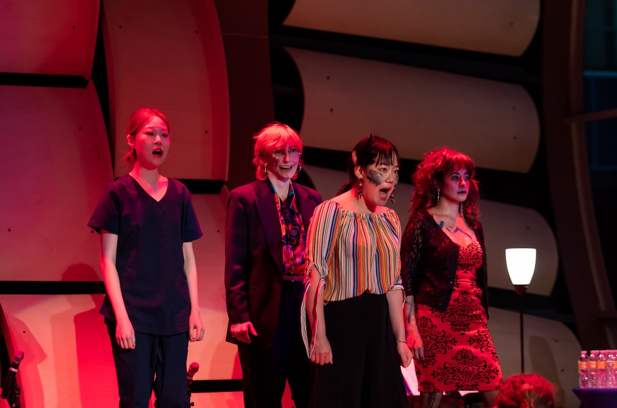 Four performers stand in a line on a stage, singing or shouting.