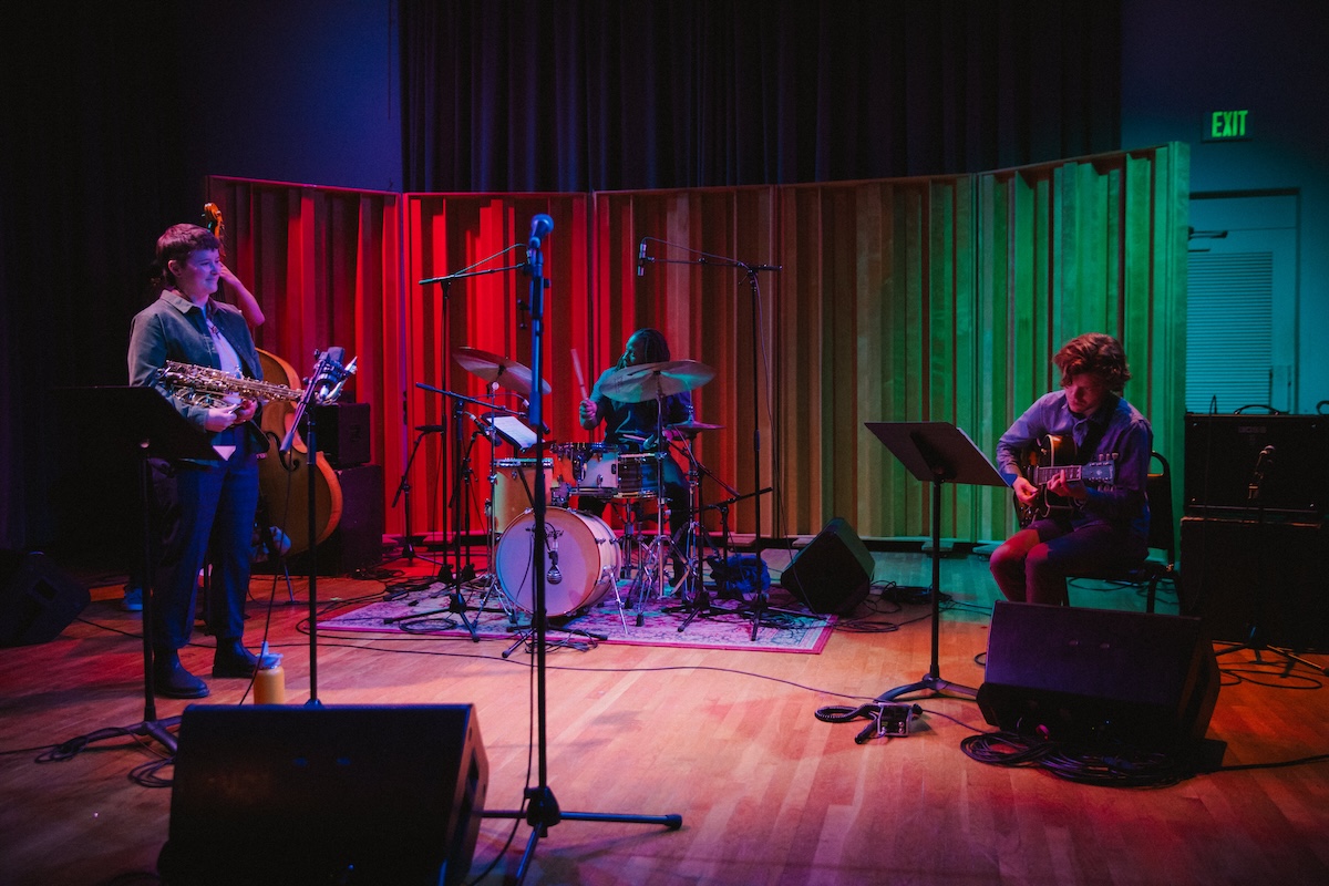 Three musicians play saxophone drums, and guitar on a small stage with colorful lights