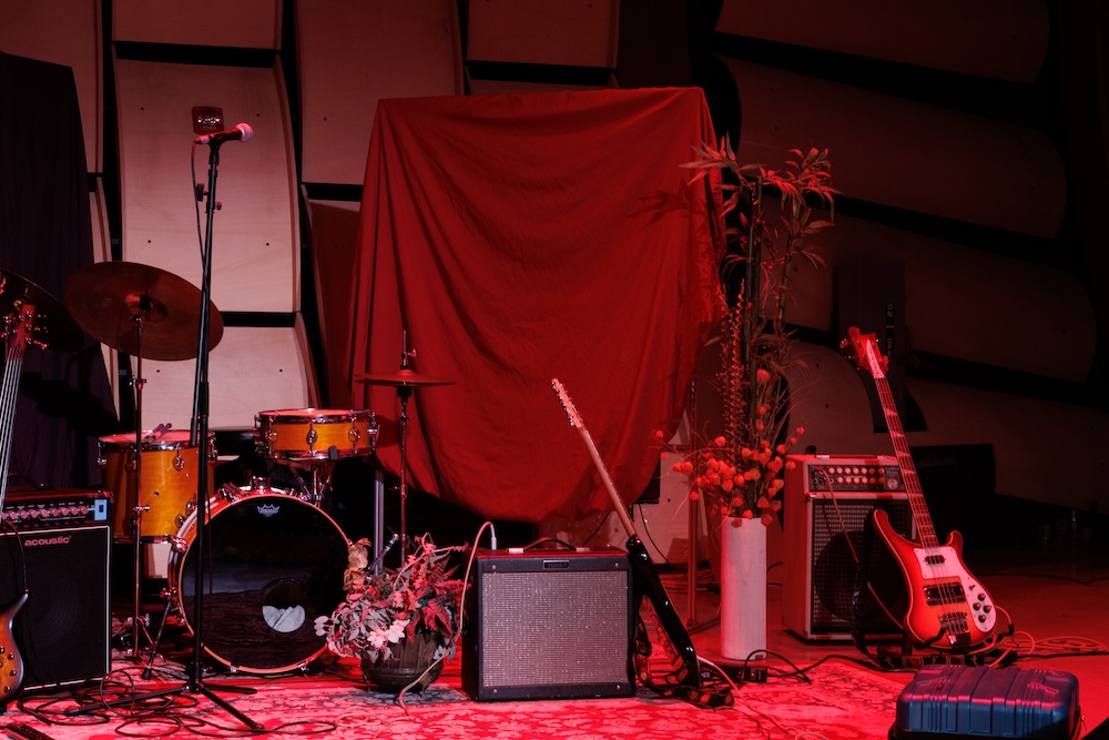 Stage setup with drum set, microphones, guitar and bass amplifiers, and guitars in front of soundproof panels.