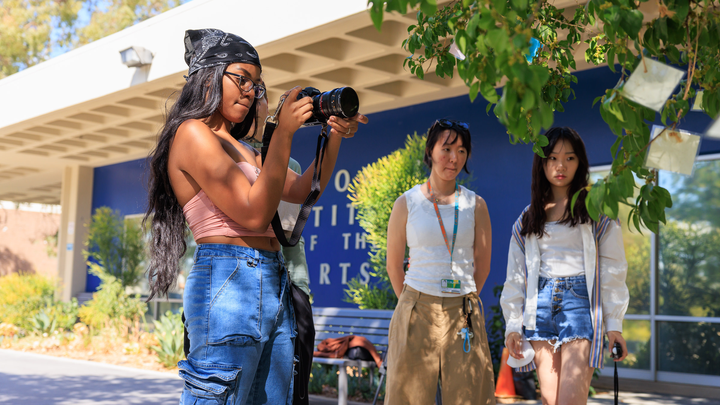 A student points a camera toward a tree as two others look on outdoors on a sunny day in front of a blue wall 