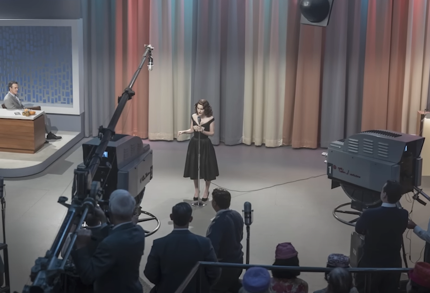 A retro television set with several cameras pointed at a lone woman in a 1950s-style dress standing and speaking into a microphone. 