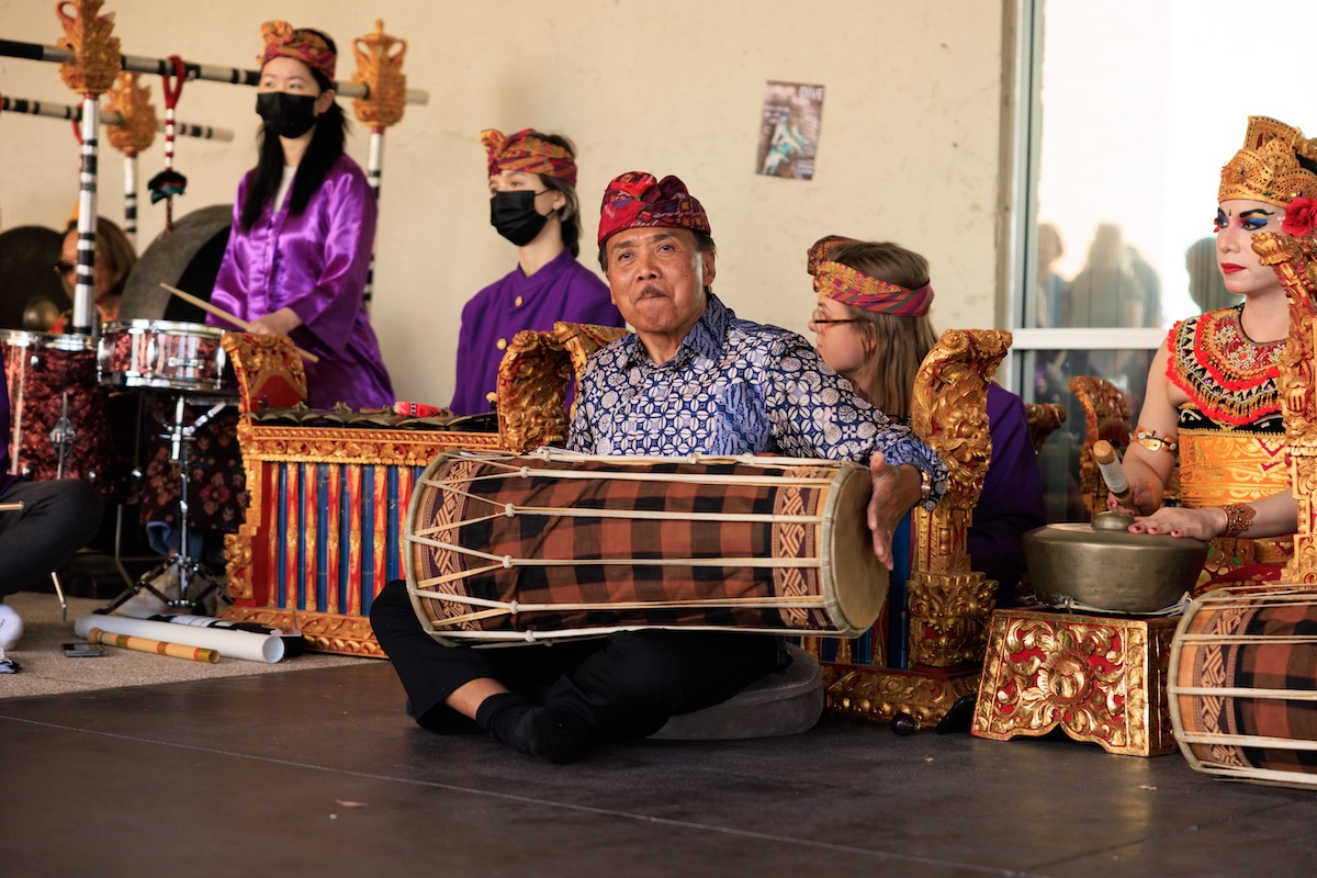 A man sits cross-legged on the floor playing a Balinese drum while other musicians play the gamelan behind him.