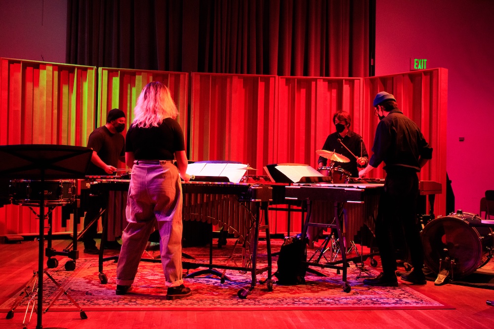 A group of four musicians stand facing each other in a circle on a red-lit stage, playing a marimba, cymbals, and other percussion instruments