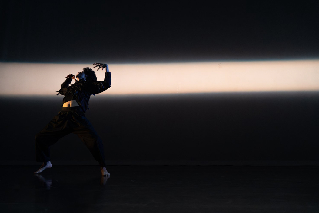 A lone dancer moves on an empty stage, in darkness except for a single bar of white light across the background