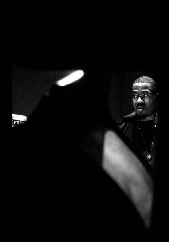 A moody, black-and-white photo of James Brandon Lewis, wearing glasses, nearly obscured by abstract dark shapes. 