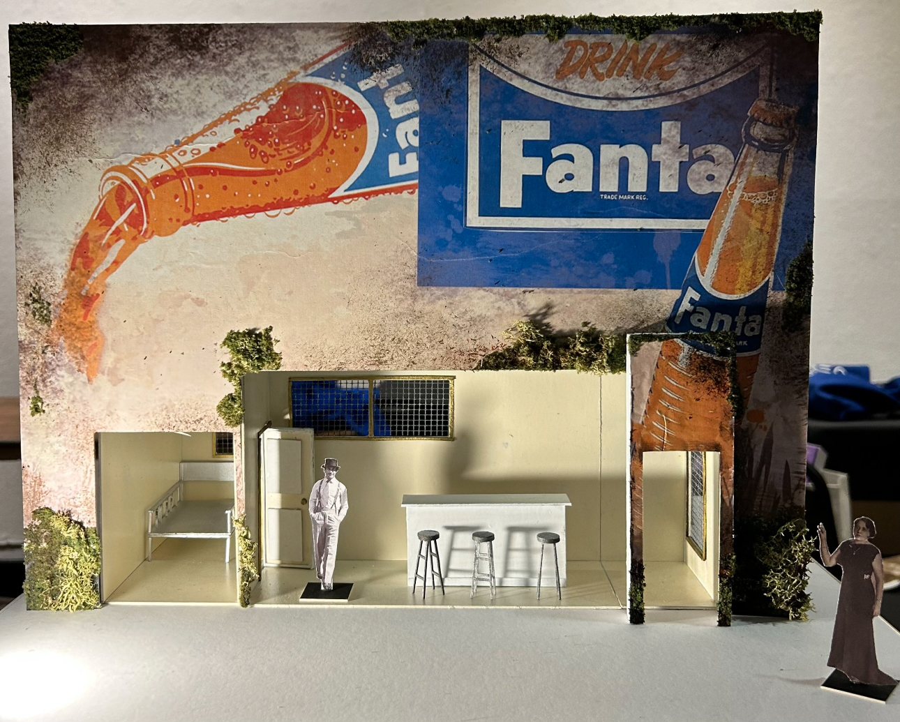 A small cardboard maquette of a kitchen or bar with two old-fashion-looking figures standing about. The exterior wall is a large, retro ad for Fanta soda.