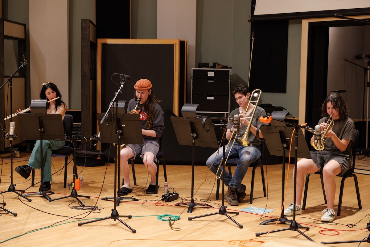 Four people sit on folding chairs in a recording studio playing wind and brass instruments. They’re surrounded by music stands, cords, and equipment.