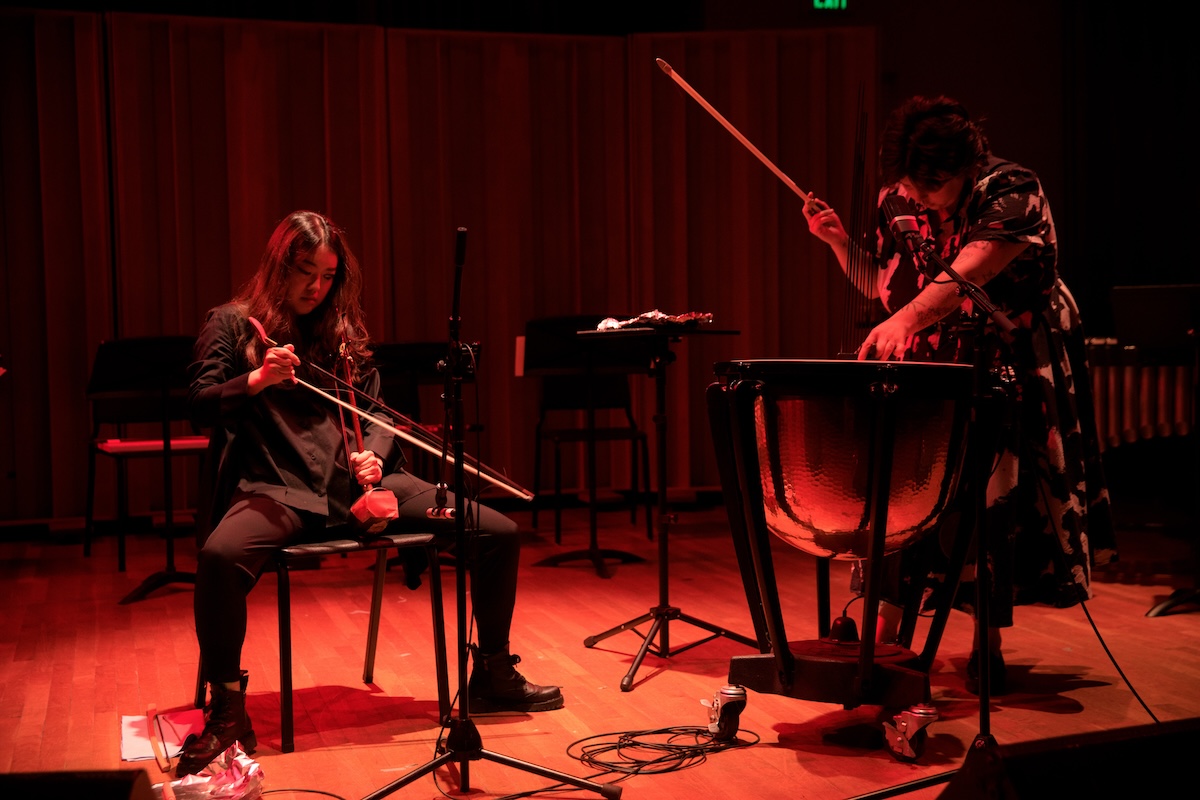 A woman dressed in black sits on a chair playing an obscure stringed instrument with a bow while another, to her right, plays a timpani drum on a stage washed in orange light.