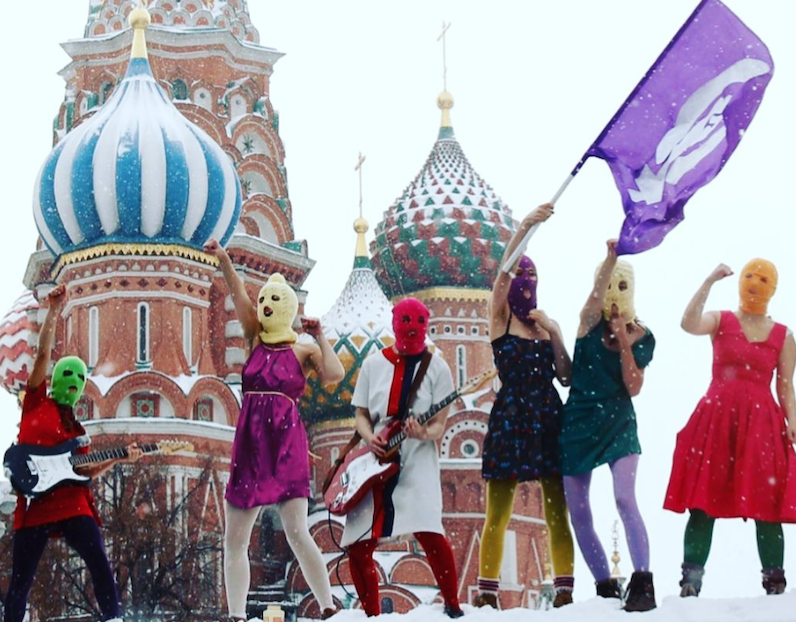 Six women in dresses and ski masks, members of Pussy Riot, stand in a line in the snow in front of the Kremlin. One waves a purple flag and two play guitars as several make power gestures with their fists.