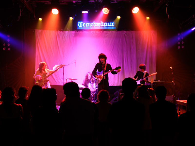 A four-person band plays on a pink-lit stage in a nightclub. The name of the club, Troubadour, is spelled out in blue neon behind them.