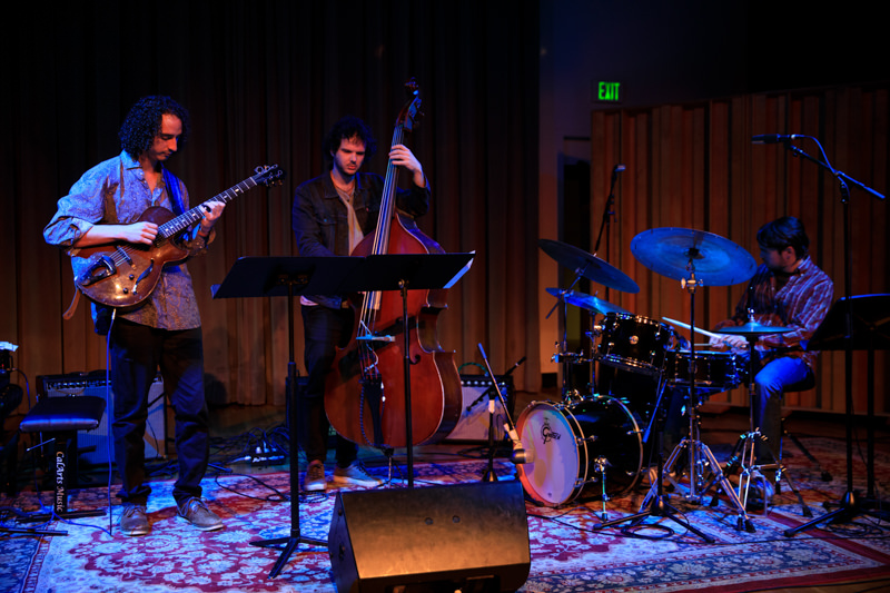 A trio of musicians play guitar, bass, and drums on a small stage