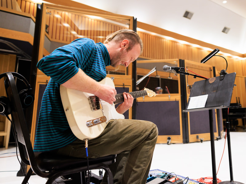 A man is sitting in a chair playing a guitar in a large institutional space. He is smiling.