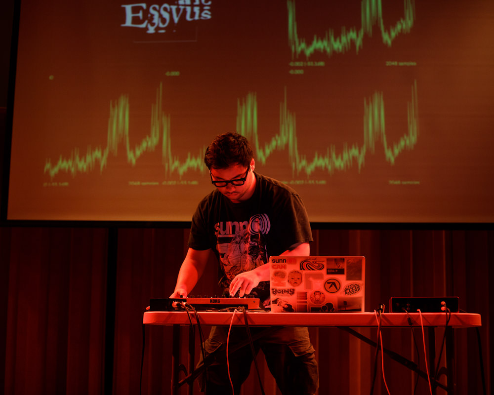 A person stands alone on a stage at a table with sound-mixing equipment. Frequency graphics are displayed on a screen behind them.