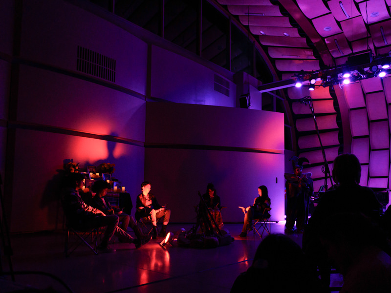 A small group of seated musicians plays on a shadowy, dramatically lit, modernistic stage.