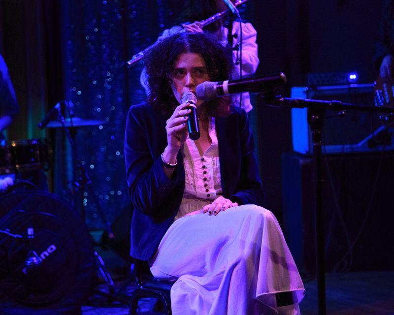 A person is seated on a blue-lit stage, singing into a microphone they’re holding and one on a stand. Someone plays the flute in the background. 