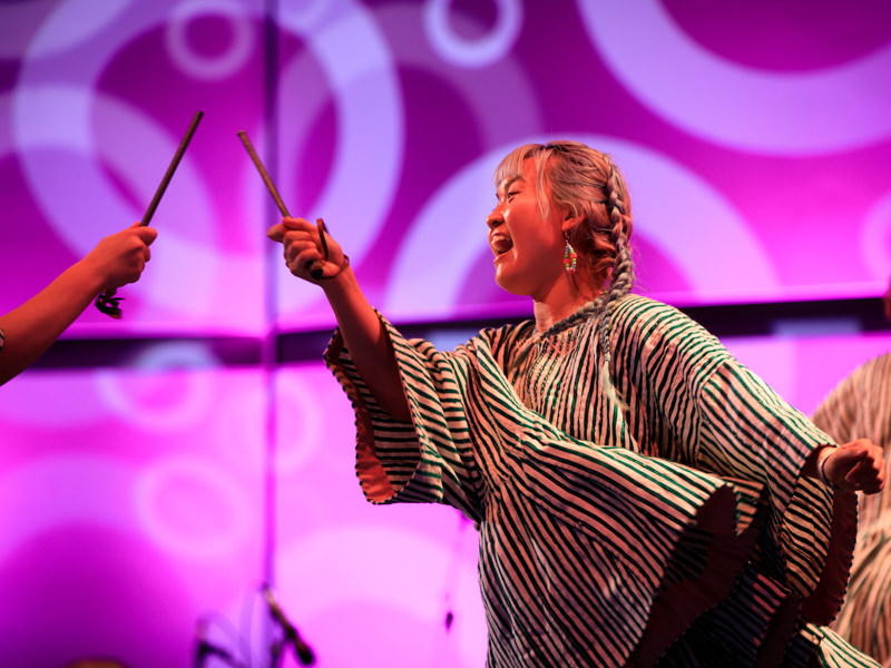 A woman in a striped outfit shouts while holding a stick up as if sword fighting with another, similar stick held by someone outside the frame. They are on a stage with a vivid pink and purple background.