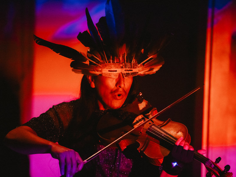 Musician wearing a feathered headpiece plays a violin under colorful ambient lighting.