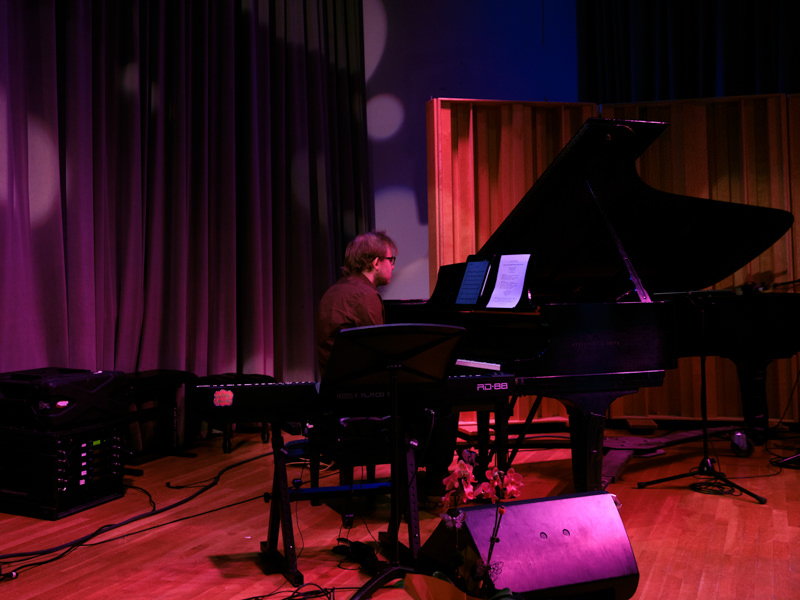 A person plays a black grand piano alone on a stage, surrounded by various audio equipment and music stands.