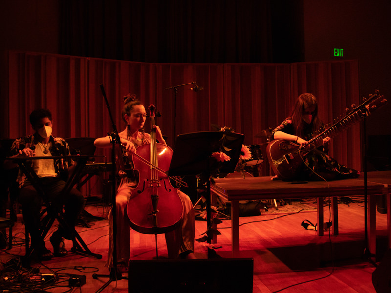 A band with a cello and a sitar perform on a stage in red light.