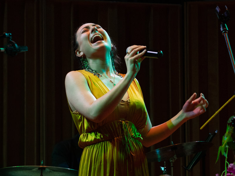 A person in a yellow dress sings dramatically holding a microphone