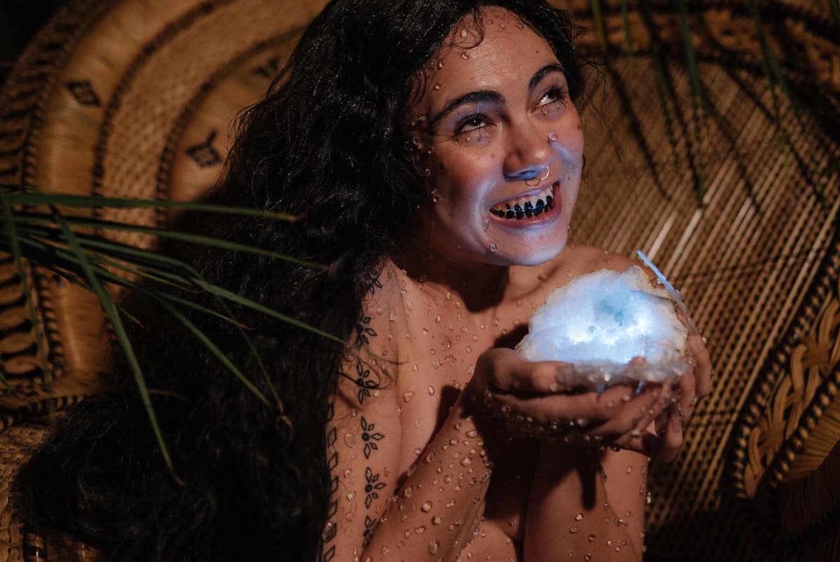 A woman with long dark hair grins, she is holding something that looks like a glowing cloud and is covered in water droplets. Her teeth are covered in black marks. 