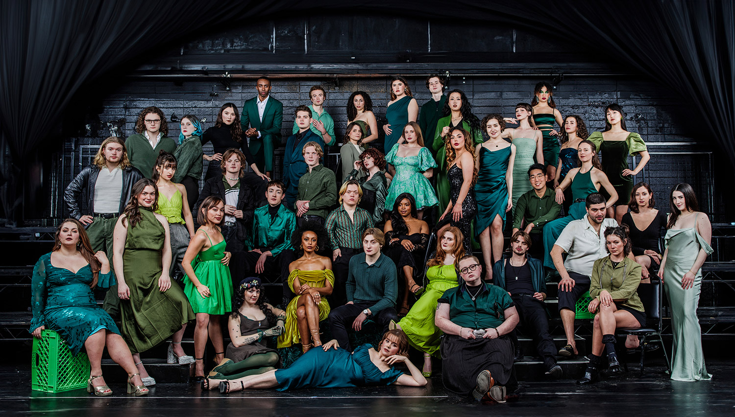 A large group of actors, dressed in glamorous clothing in shades of green, poses on a theater stage