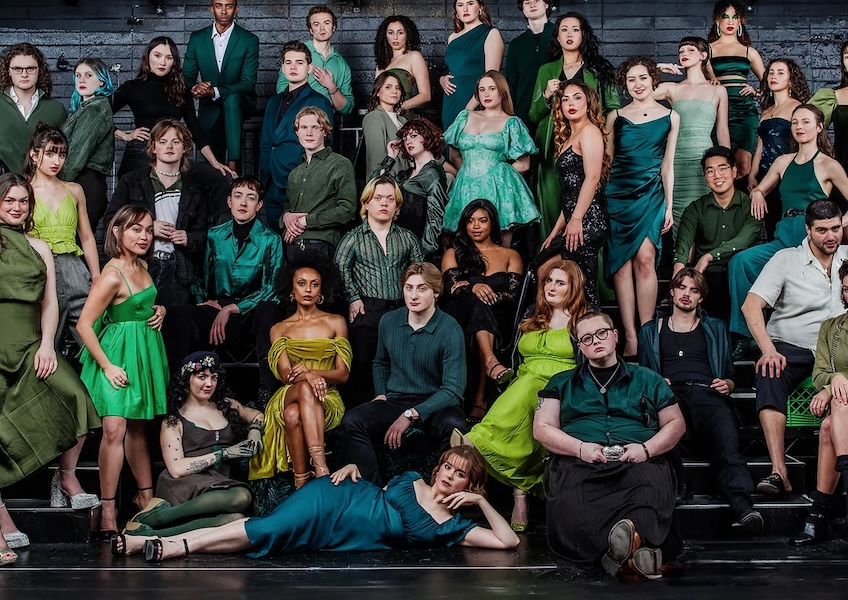 A large group of actors, dressed in glamorous clothing in shades of green, poses on a theater stage