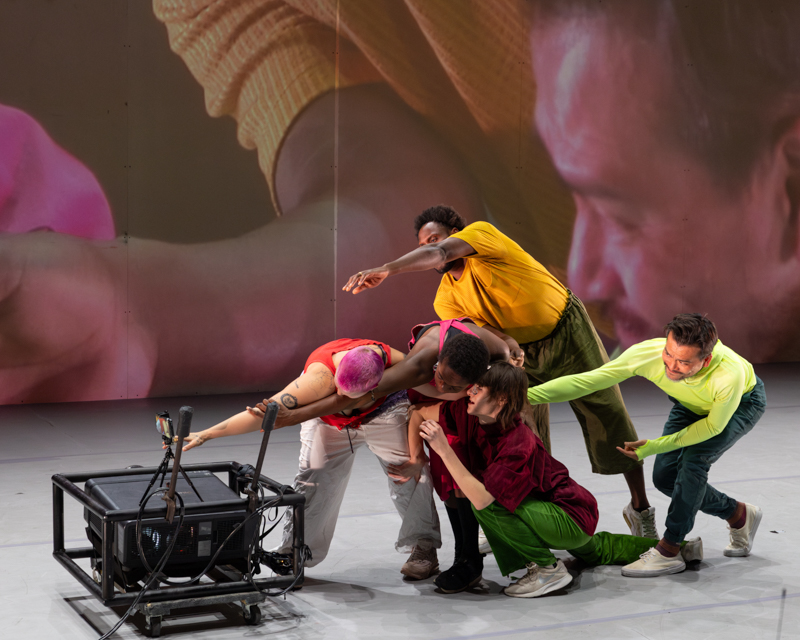 A group of actors, wearing bright colored clothes, reach toward a black box. The stage behind them is a projected image. 