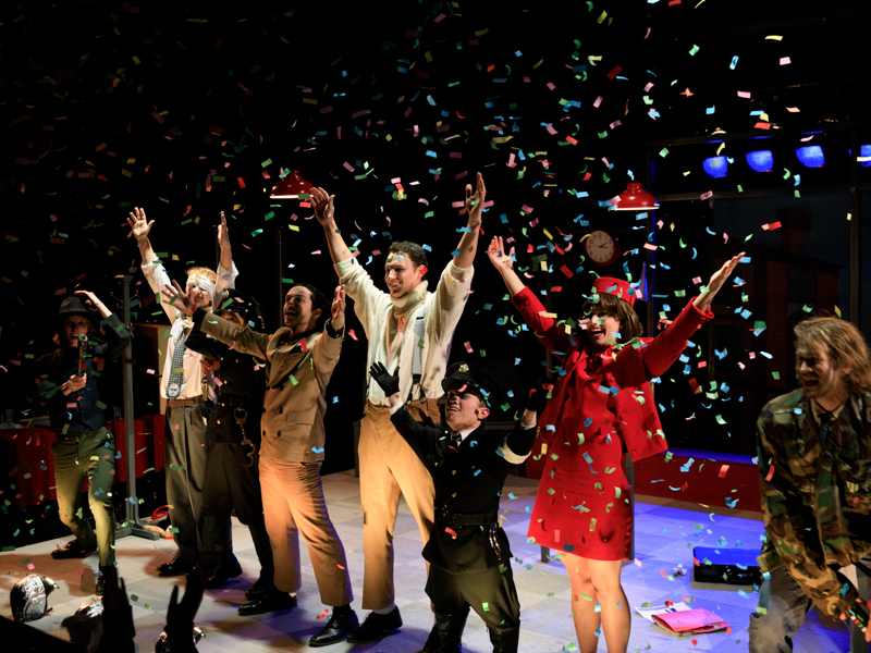 A line of actors standing on a stage with their arms and hands raised as confetti falls around them