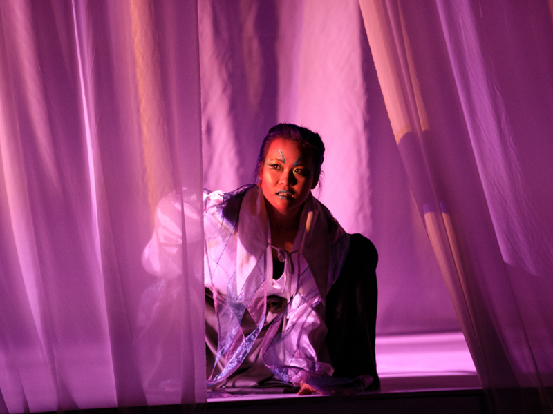 Woman in dramatic makeup crouching among theatrically lit pink curtains on a stage