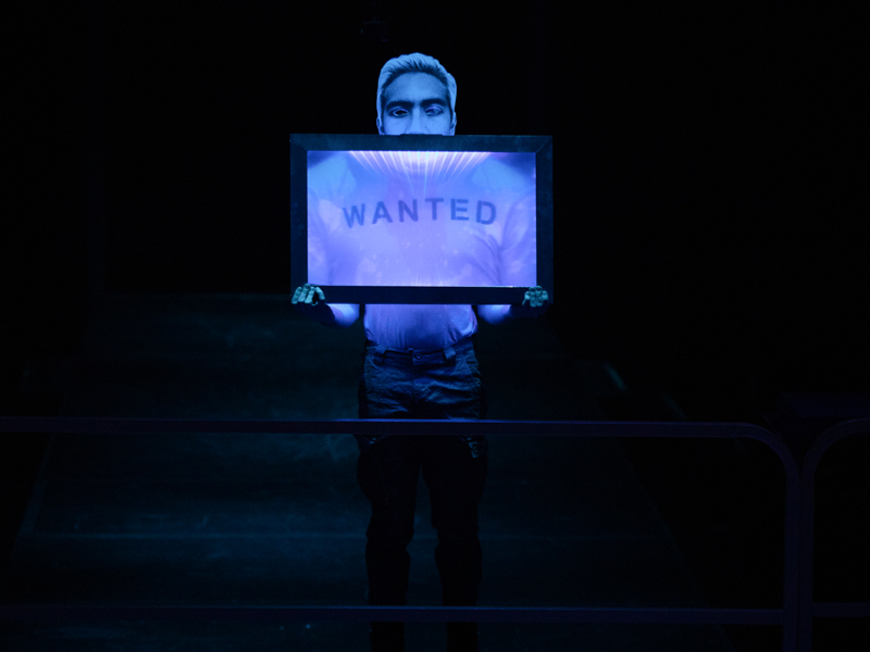 Person surrounded by darkness and lit with blue and purple light holding a sign that says “wanted.”