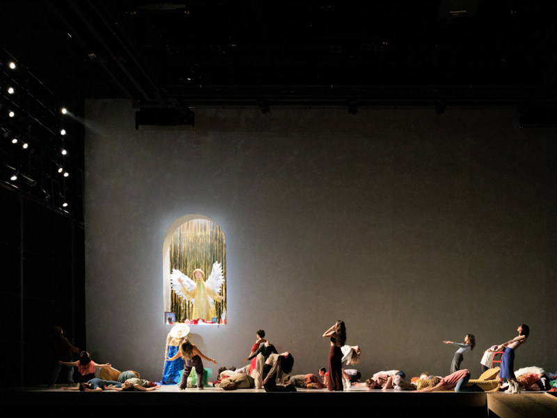 Seen from a distance in a theater, a group of performers on stage faces a nook featuring a sculpture of an angel