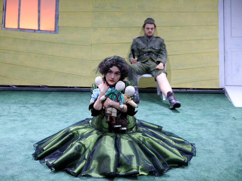 A woman sitting on a stage holding three dolls as a man sits behind her