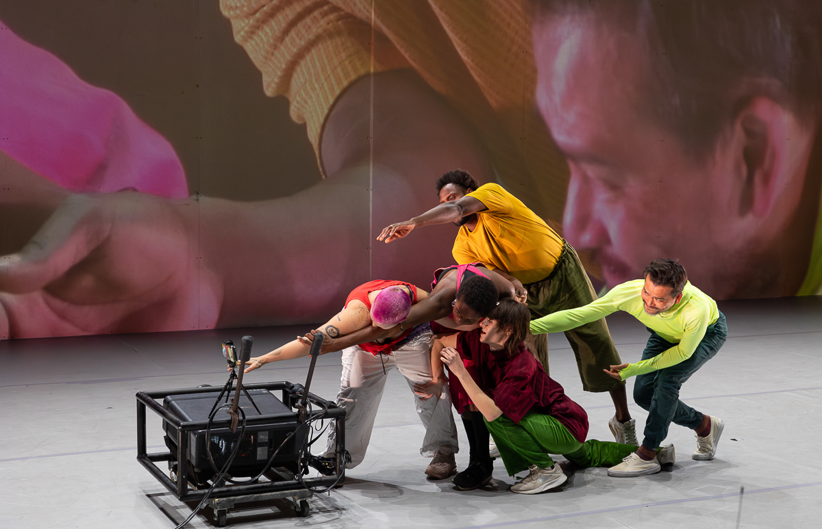 A group of performers on stage posing in front of a camera as their image is projected behind them