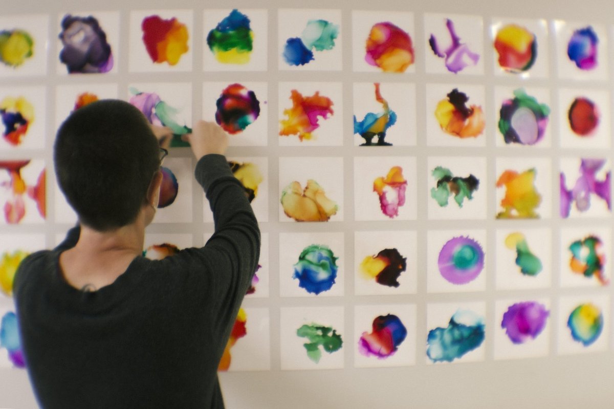 Person pinning small, colorful, abstract paintings to the wall in a grid pattern. 