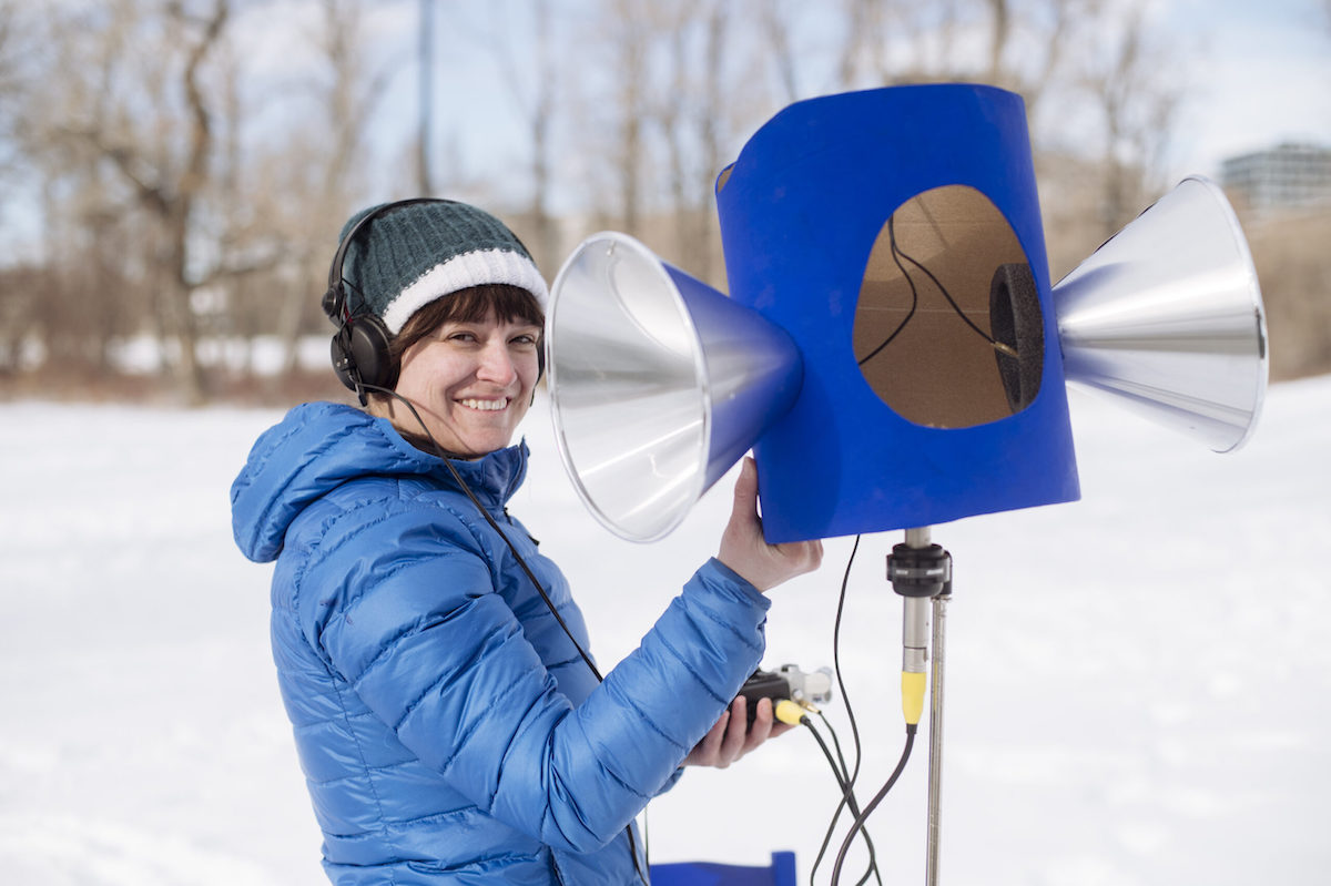 A woman wearing a blue parka, warm hat, and headphones stands next to a blue and silver contraption that resembles a megaphone on a tripod. The scene is outdoors in the snow on a sunny day. 